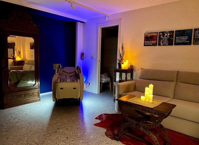 A room with a massage chair, a sofa, a table with candles, and wall art. 1