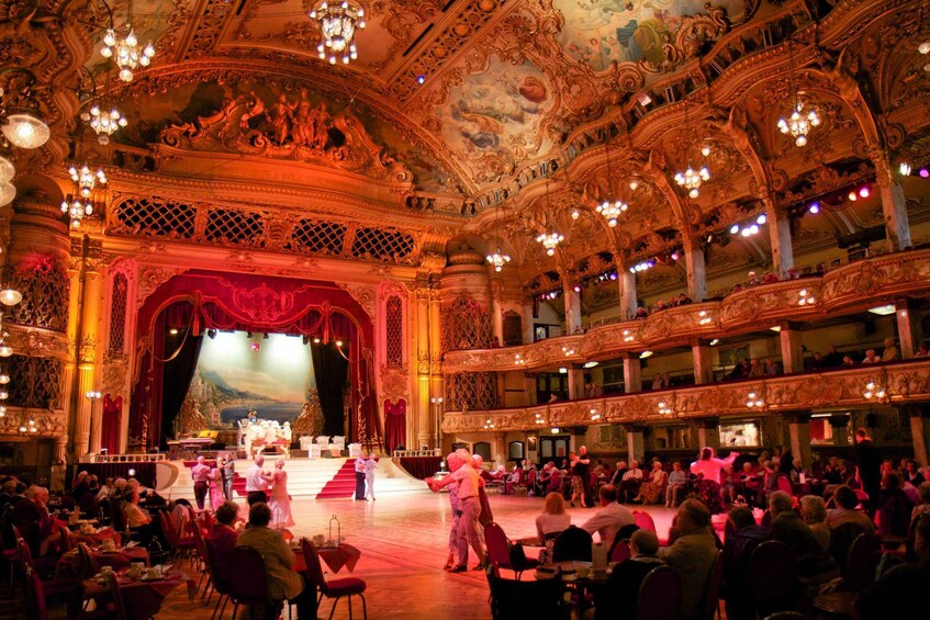 An ornate theater with a red stage and audience seated in rows. 1