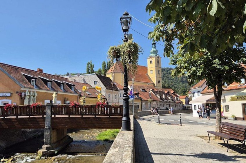 A riverside walkway with a wooden bridge, a street lamp, and buildings with red-tiled roofs. 1