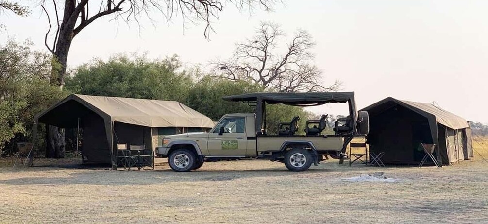 A beige off-road vehicle parked between two safari tents in a dry, grassy area with sparse trees in the background. 3