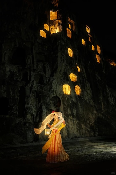 A person in traditional attire performing a dance in front of a cave with illuminated windows. 4