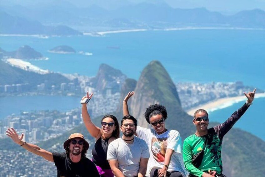 A group of people posing with arms raised against a scenic backdrop of a coastal city and a prominent mountain peak. 5