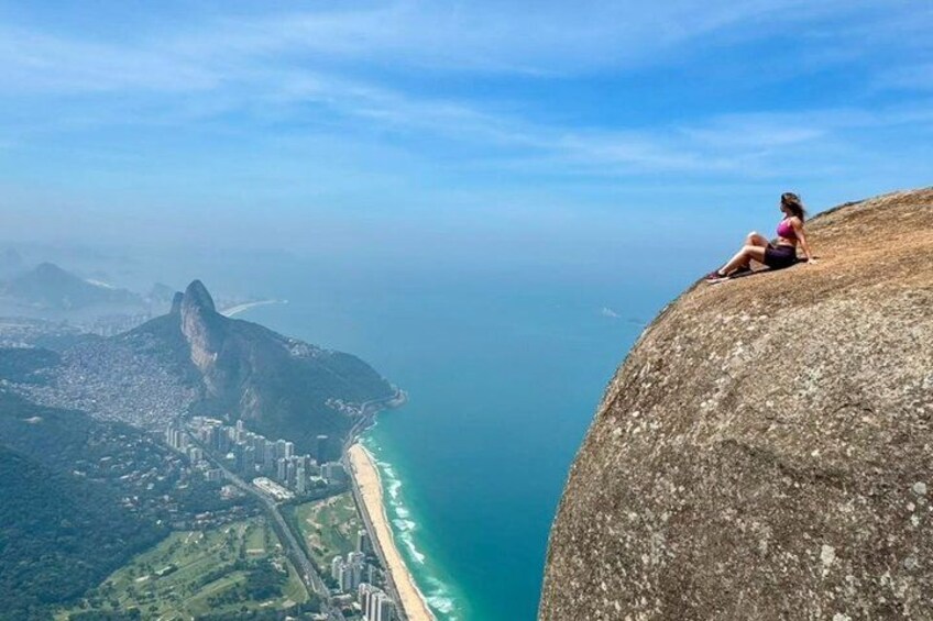 A person sitting on a cliff overlooking a coastal city with a prominent rock formation. 1