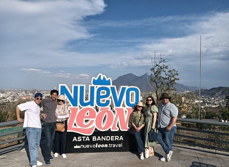 A group of people standing in front of a sign with 'Nuevo Leon' and 'Asta Bandera' written on it, with a cityscape and mountains in the background. 5