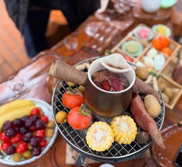 A traditional food setup with a steaming pot, corn on the cob, sweet potatoes, and a variety of fruits on a metal tray. 1