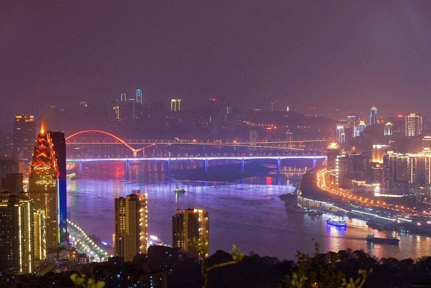A cityscape at night with illuminated buildings, a river, and a bridge with colorful lights. 3