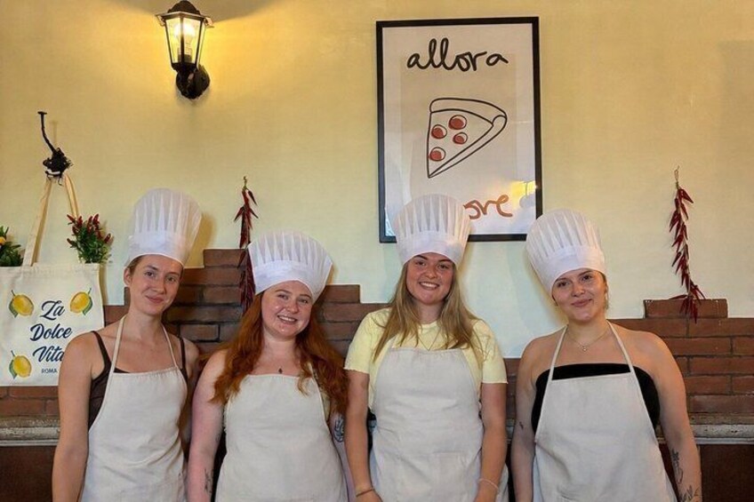 Four people in chef hats and aprons standing in front of a wall with a pizza slice and the word 'allora' on it. 4