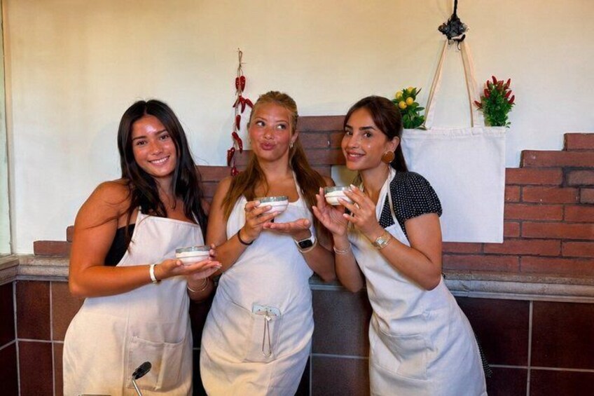 Three individuals in aprons holding bowls, standing in a kitchen with a brick wall and a hanging plant in the background. 5