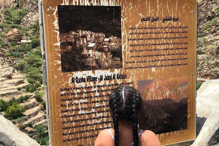 A weathered informational sign with text and a photograph of Al Gasha Village. 2