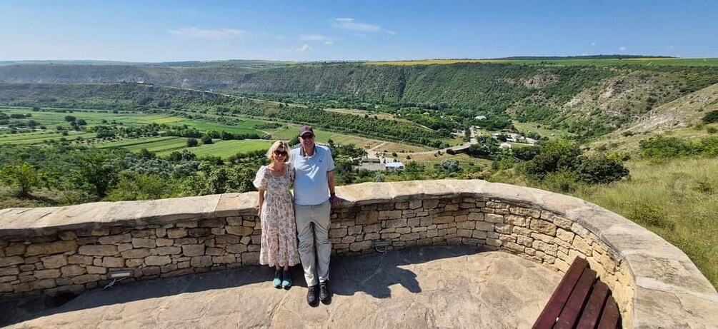 A couple standing on a stone bench overlooking a valley with fields and a distant village. 1