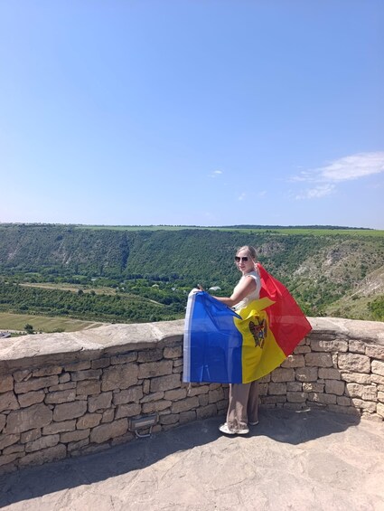 A person holding a flag with a blue and yellow emblem, standing on a stone wall overlooking a hilly landscape. 2