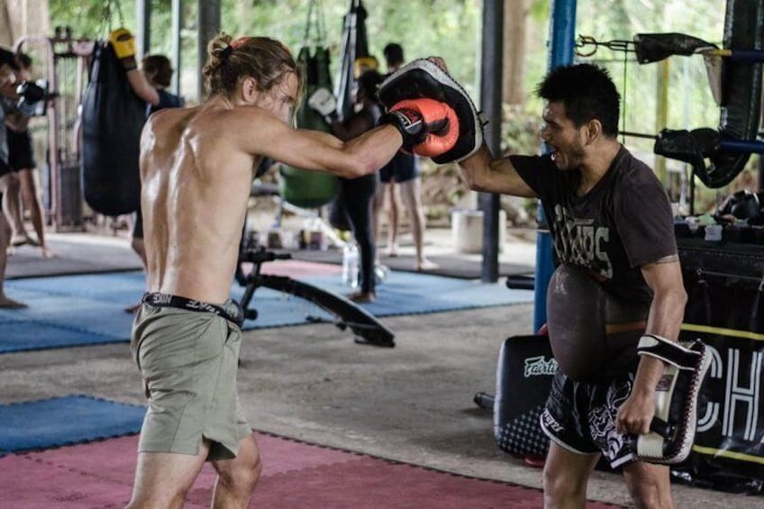 Two people in a gym, one wearing boxing gloves and the other holding a punching bag. 1