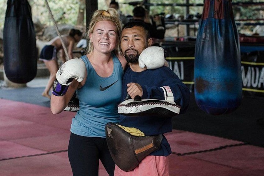 A person in a blue tank top and boxing gloves, another in a blue jacket with a white headband, and a punching bag in a gym. 2