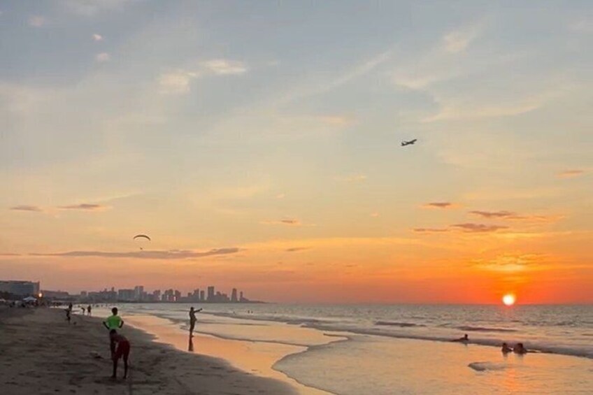 A beach at sunset with a city skyline in the background, a person walking, and a kite flying. 2