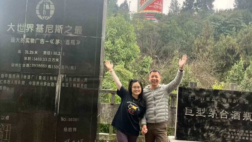 Two people standing in front of a large black monument with inscriptions, surrounded by greenery. 3