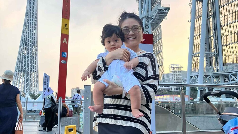 A woman holding a baby in an urban setting with tall structures and signage. 1