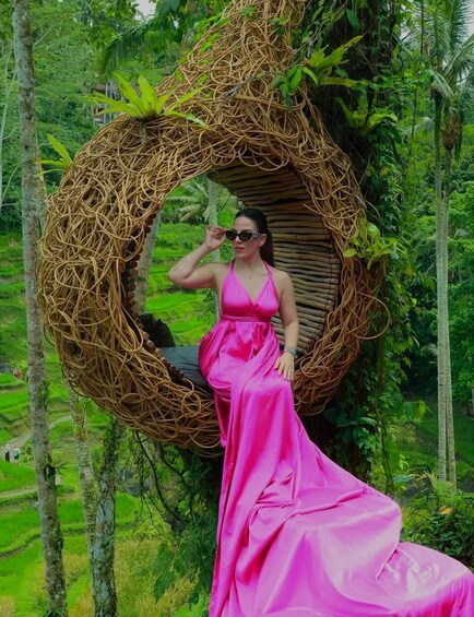 A woman in a pink dress sitting on a circular wicker structure surrounded by lush greenery. 3