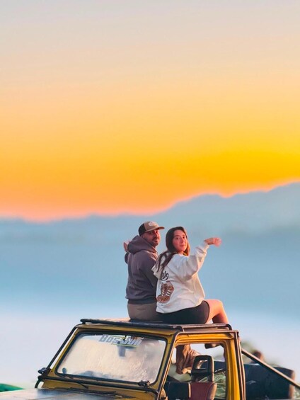Two people standing on the roof of a yellow vehicle with a sunset in the background. 1