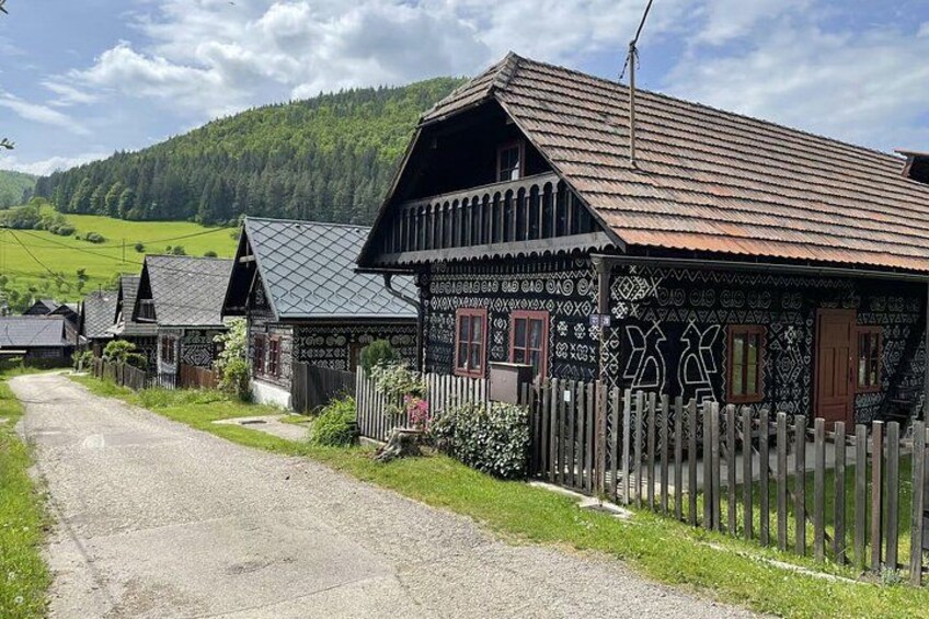 A row of traditional wooden houses with patterned facades, a gravel road, and a green hillside in the background. 2