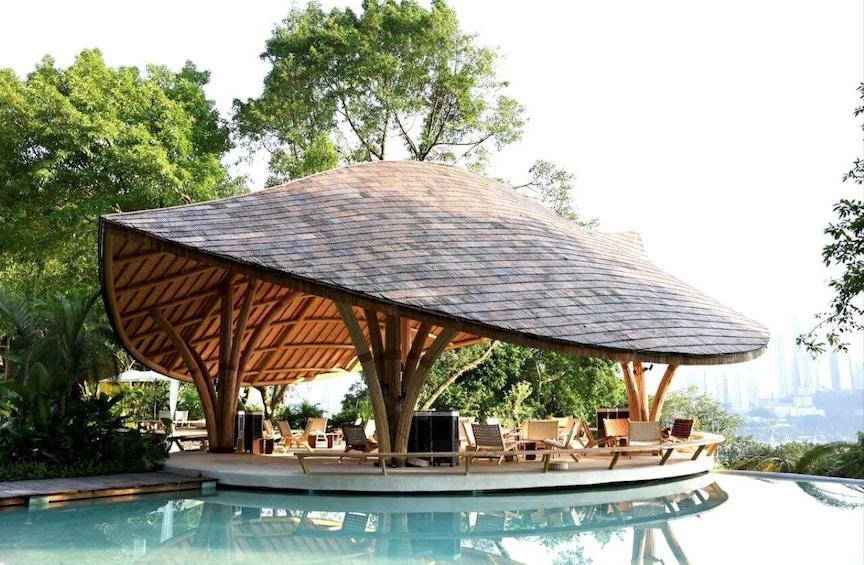 A poolside lounge area with a thatched roof structure, surrounded by lush greenery and overlooking a cityscape. 1