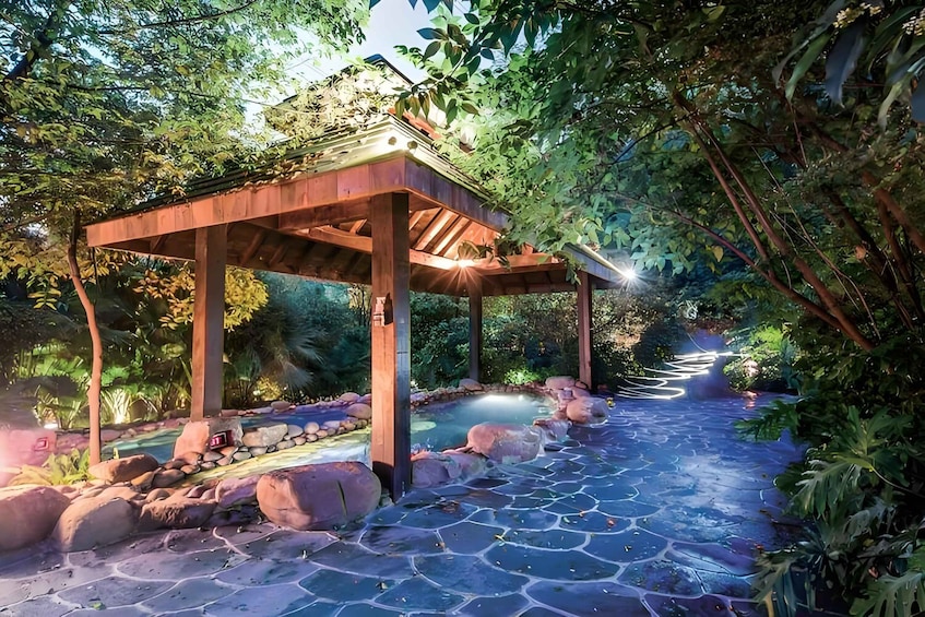 A wooden gazebo with a stone pathway leading to a pond, surrounded by lush greenery. 3