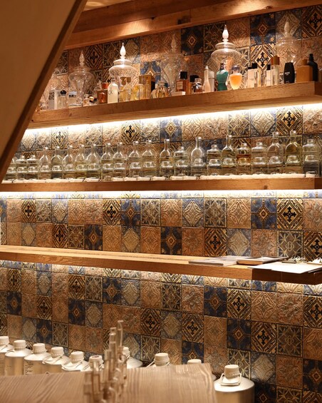 A shelf with various glass bottles and containers, some with labels, set against a backdrop of patterned tiles. 2