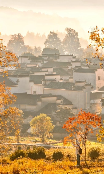 A person taking a photo of a traditional village with tiered buildings and autumn foliage. 2