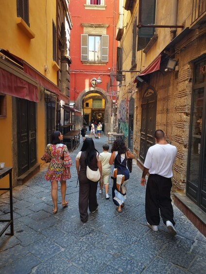 A narrow cobblestone street lined with colorful buildings, a few pedestrians, and a large archway leading to a building with a sign. 2