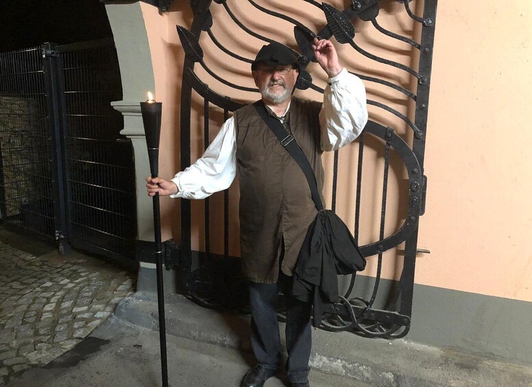 A person standing in front of a metal gate with a curved design, holding a walking stick, and wearing a vest and hat. 2