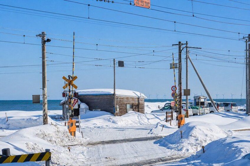 A snow-covered railway crossing with warning signs, a wooden structure, and vehicles parked nearby. 1