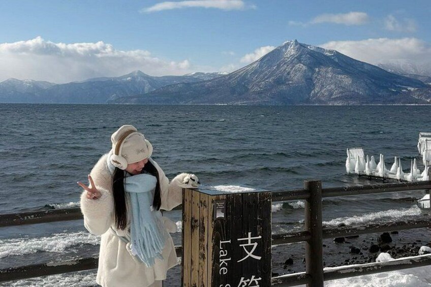 A person by a wooden sign with Japanese characters, a snowy landscape, and a body of water. 5