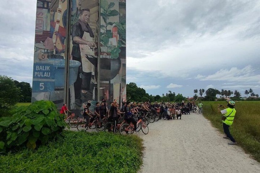 A group of cyclists on a dirt path, a large mural with cooking scenes, and a person in a high-visibility vest. 5