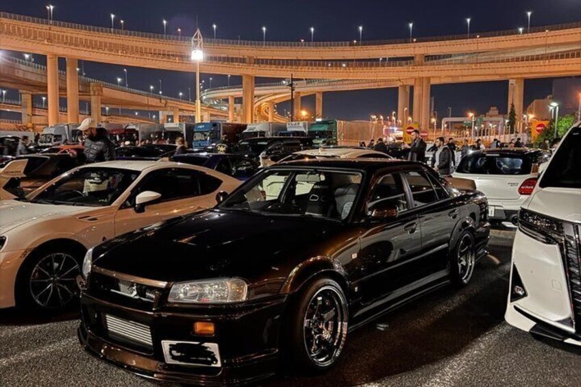 A black sports car parked in a crowded lot with a large overpass in the background. 1