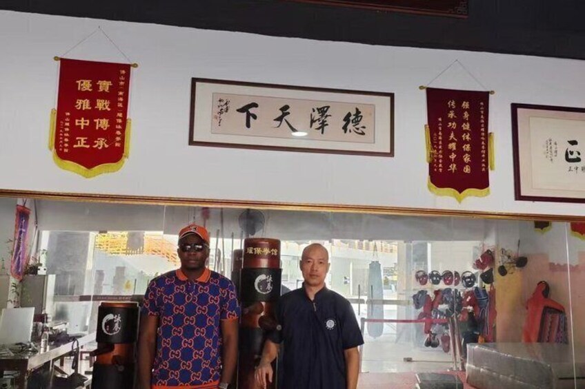 Two individuals standing in front of a shop with Chinese characters on the wall and various items displayed inside. 5