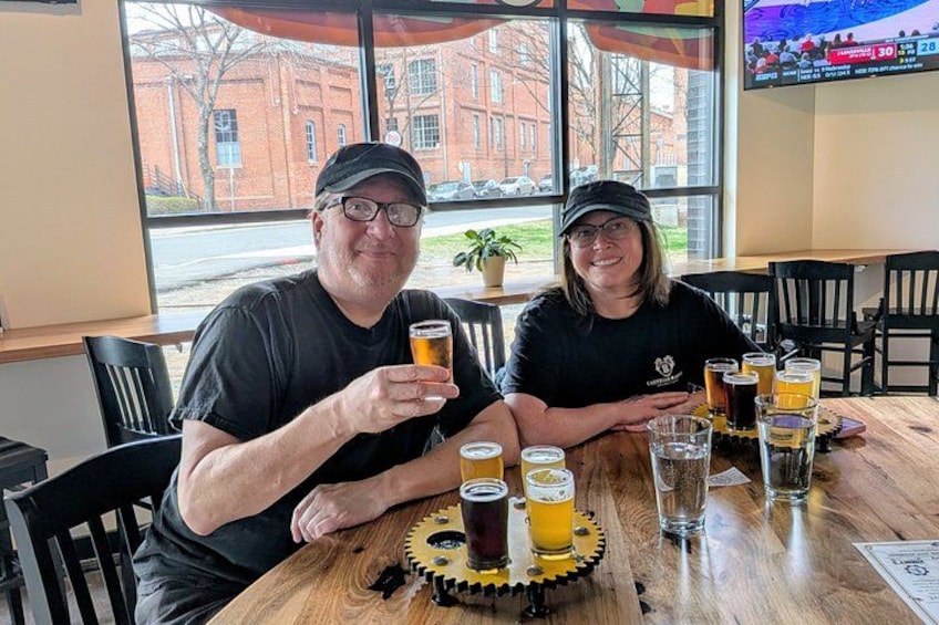Two people sitting at a table with multiple glasses of beer and a coaster with a gear design. 5