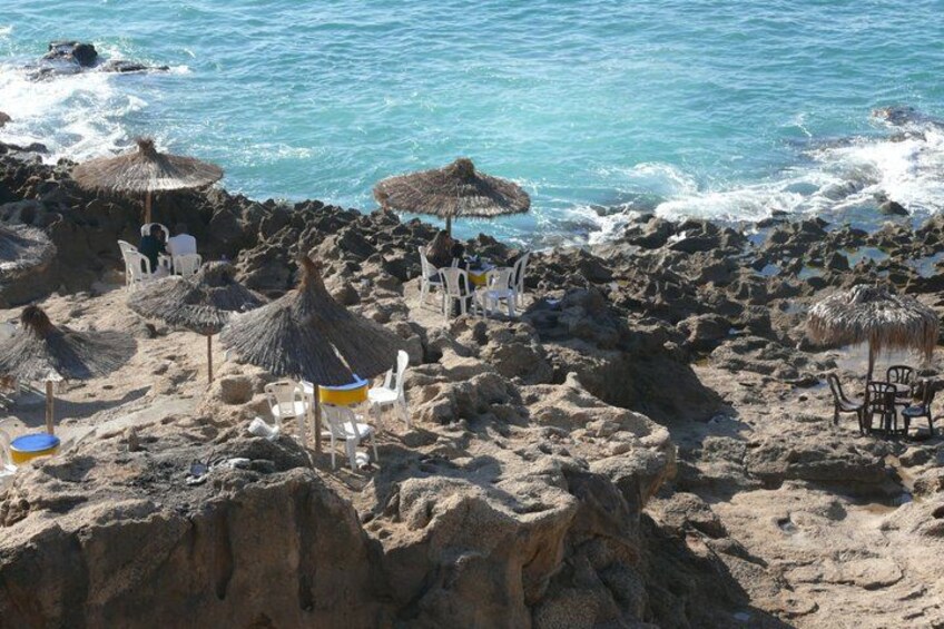 A rocky beach with thatched umbrellas and white plastic chairs. 3