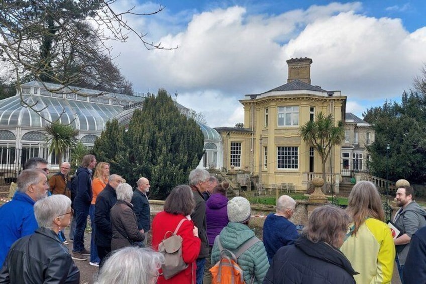 A group of people gathered in a garden with a large glass greenhouse and a historic building in the background. 3