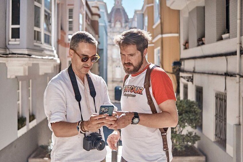 Two people standing on a street, one holding a camera and the other looking at a smartphone. 1