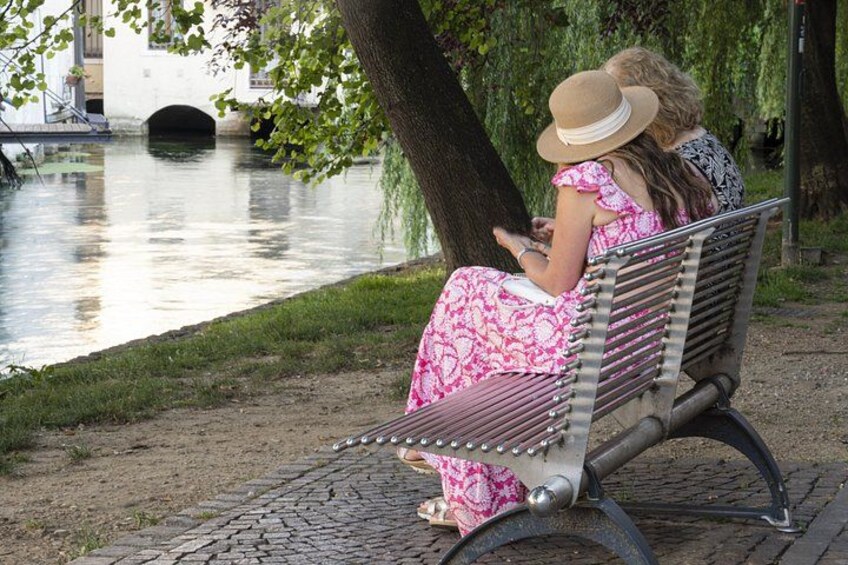 A woman in a pink dress and hat sitting on a bench by a river. 3