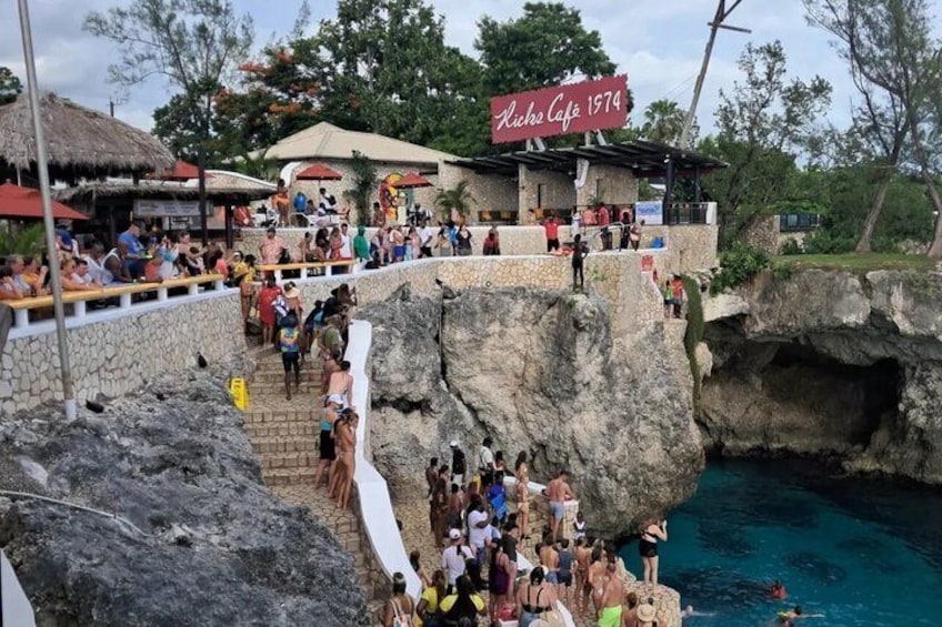 A rocky cliffside with a crowd of people, a sign reading 'Rico's Cafe 1974', and a pool below. 5