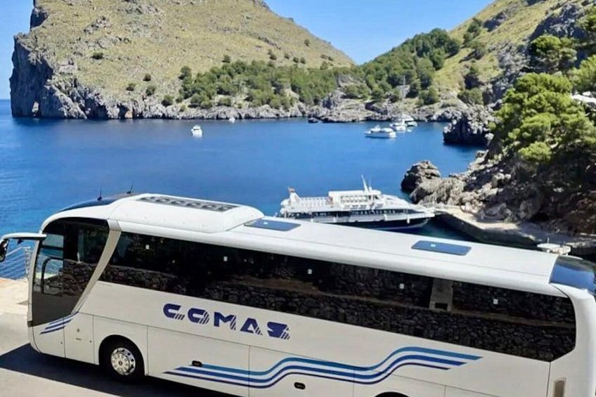 A white bus with blue stripes parked near a body of water with boats and rocky terrain in the background. 5