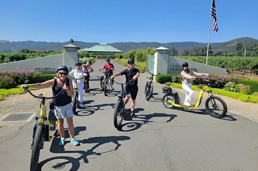 A group of people riding bicycles and a scooter on a paved path with a sign that reads 'LAIRD FAMILY' and a flag in the background. 2