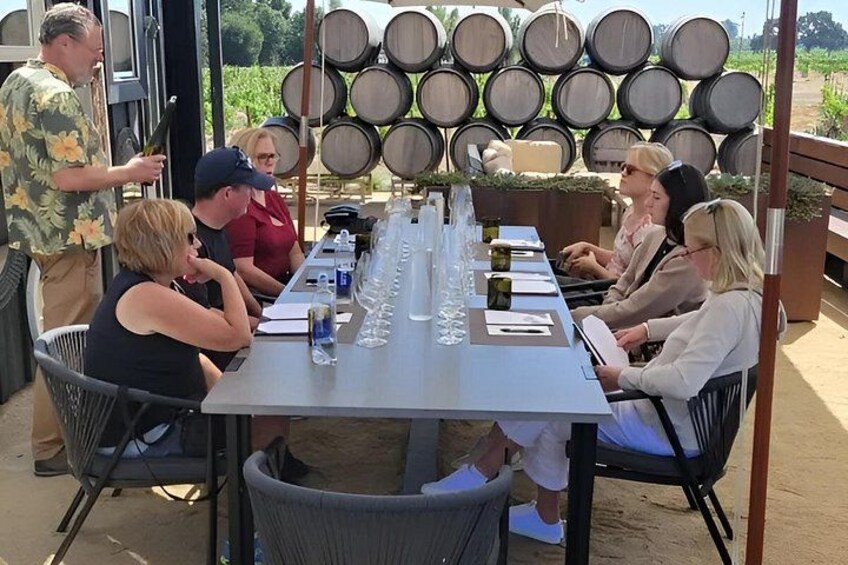 A group of people seated at a table with wine glasses and bottles, outdoors with barrels in the background. 3