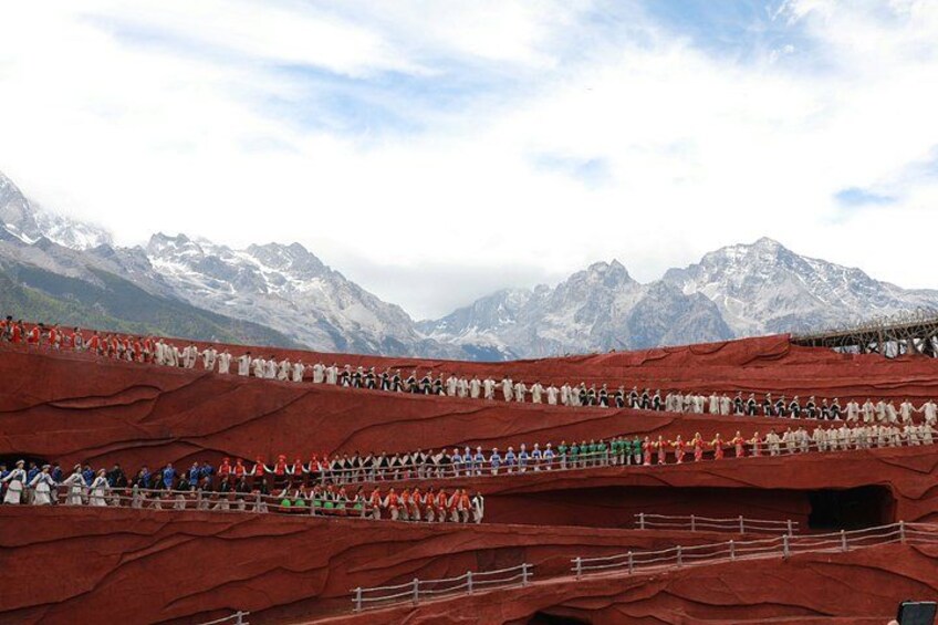 A large outdoor event with people seated on tiered red platforms, set against a mountainous backdrop. 4