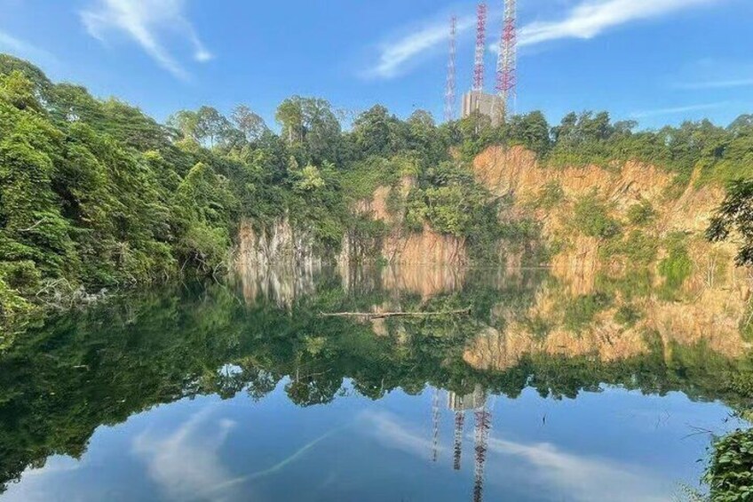 A calm lake with dense greenery and a clear reflection of the sky. In the background, there are two tall communication towers. 1