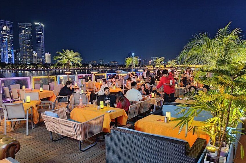 A rooftop bar with tables and chairs, palm trees, and a city skyline in the background. 5