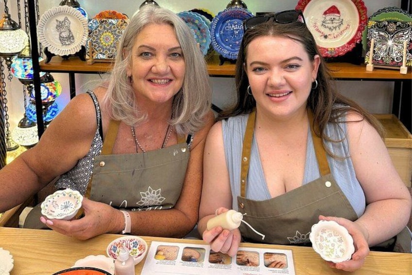 Two individuals holding ceramic bowls, with a display of colorful plates in the background. 5