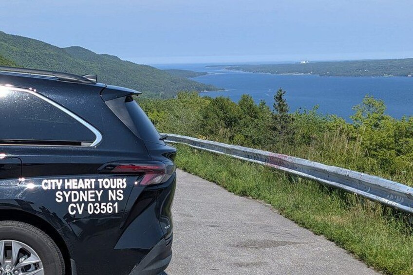 A black SUV with 'CITY HEART TOURS SYDNEY NS CV 03561' on its rear, parked on a road with a guardrail, overlooking a body of water and hills. 5