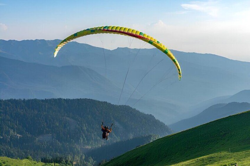 A paraglider in mid-flight over a mountainous landscape with green hills and forested areas. 3