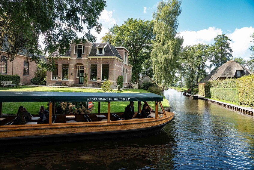 A boat with 'RESTAURANT DE RIETSTUP' on its side, a brick building, and a thatched-roof structure along a canal. 1